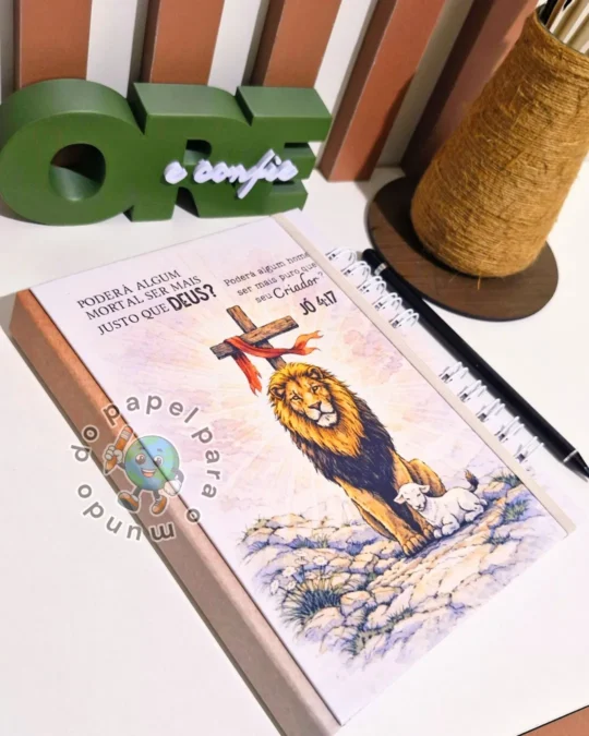 Caderno devocional com espiral preto, capa ilustrada com um leão diante de uma cruz e versículo impresso